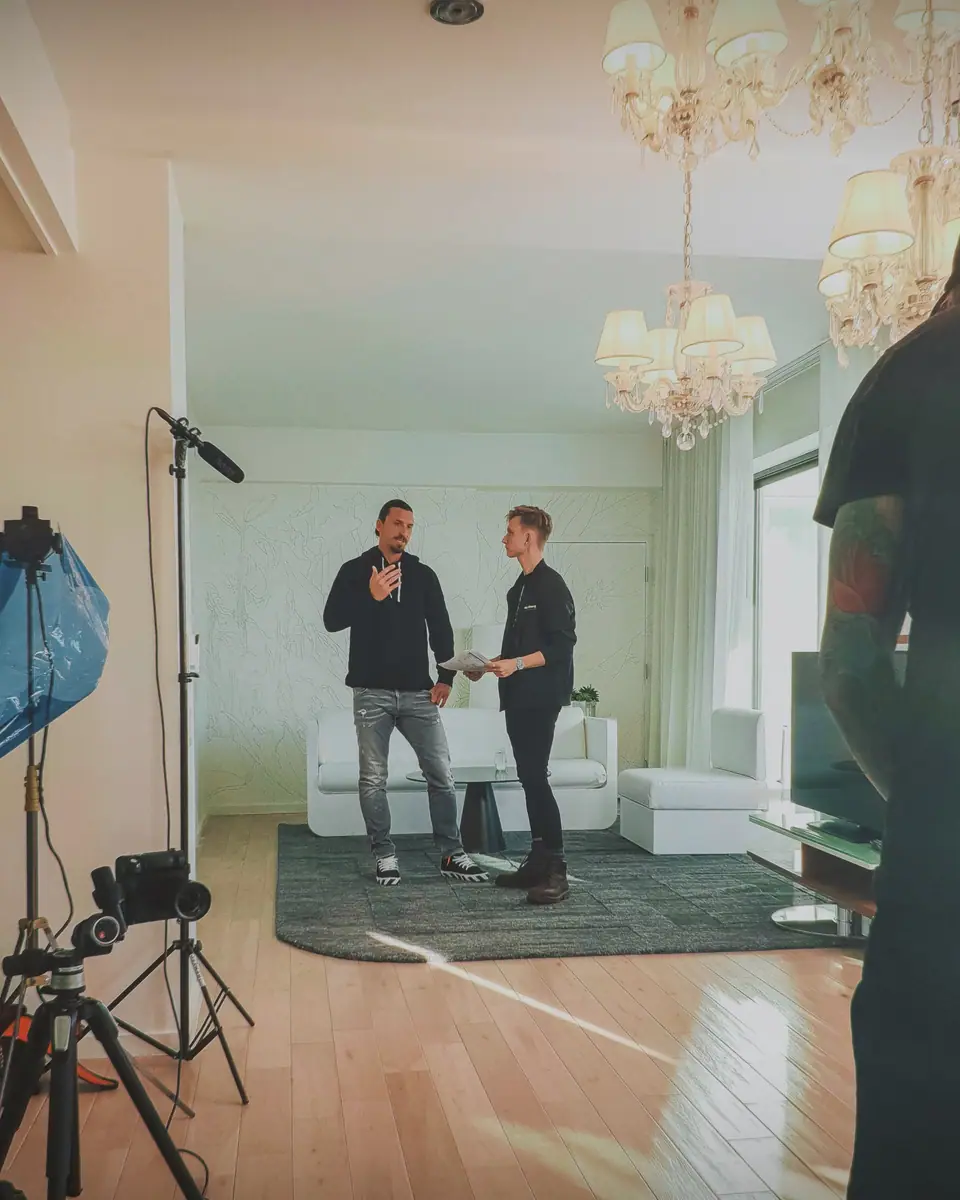 Crew filming two people in a bright apartment with chandeliers and a white sofa, a camera on a tripod in the foreground