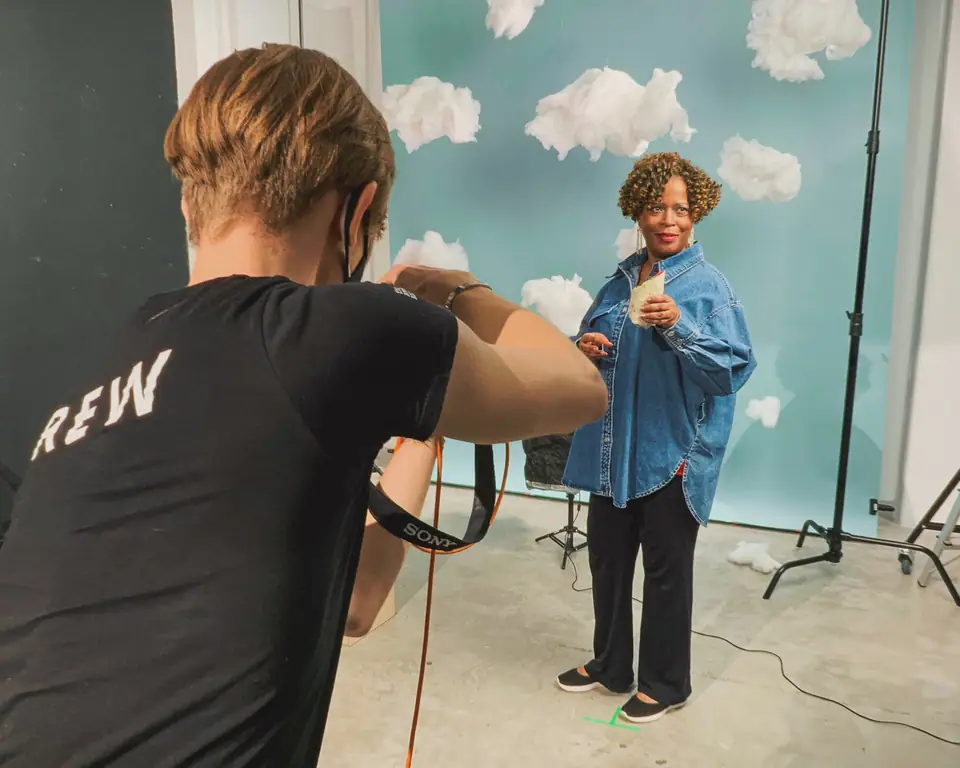 A photographer in a CREW T-shirt shooting a woman holding a drink against a blue backdrop with paper clouds