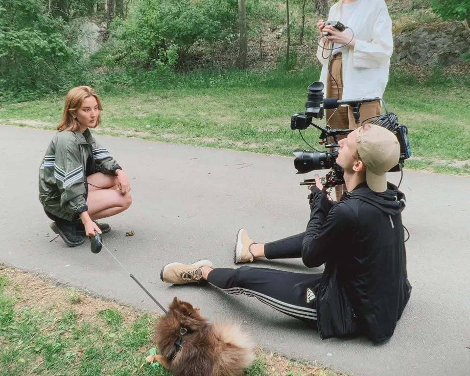 A camera operator sitting on a path filming a young woman crouching next to a small fluffy dog