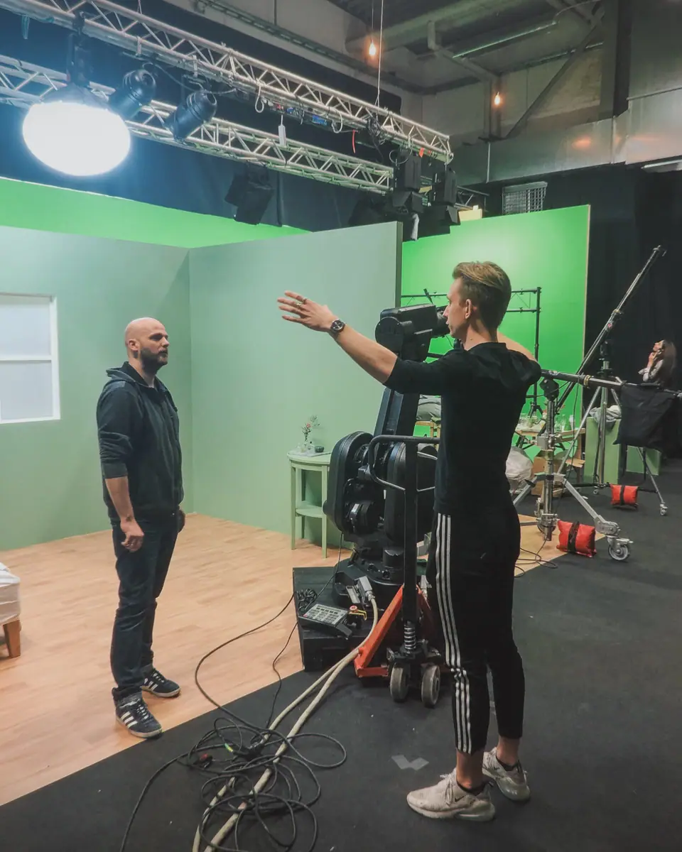 A director giving an actor directions in a green-screen studio, a camera crane beside him