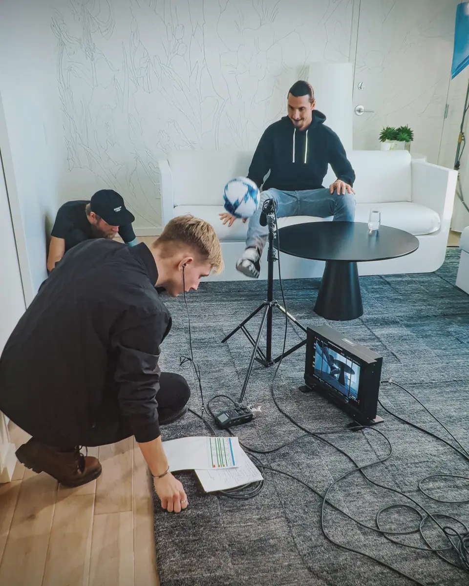 Zlatan Ibrahimović on a white sofa with a football while crew check a monitor on the floor