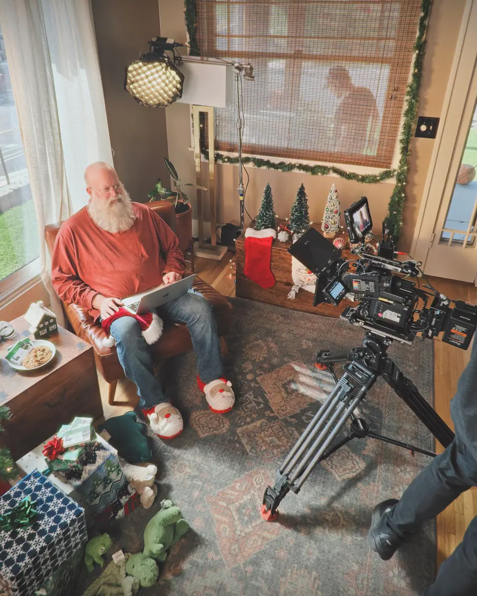 A cinema camera filming a white-bearded man in a red sweater with a laptop in a living room decorated for Christmas