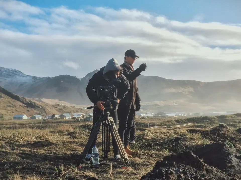 Two crew members with a camera on a tripod on a volcanic plain in Iceland, mountains behind them