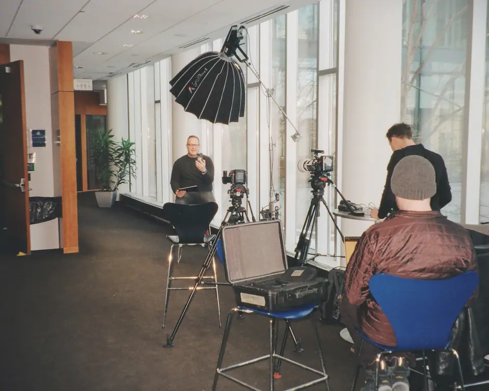 An interview set by the windows of an office, a man on a stool under a large softbox, crew and a laptop in the foreground