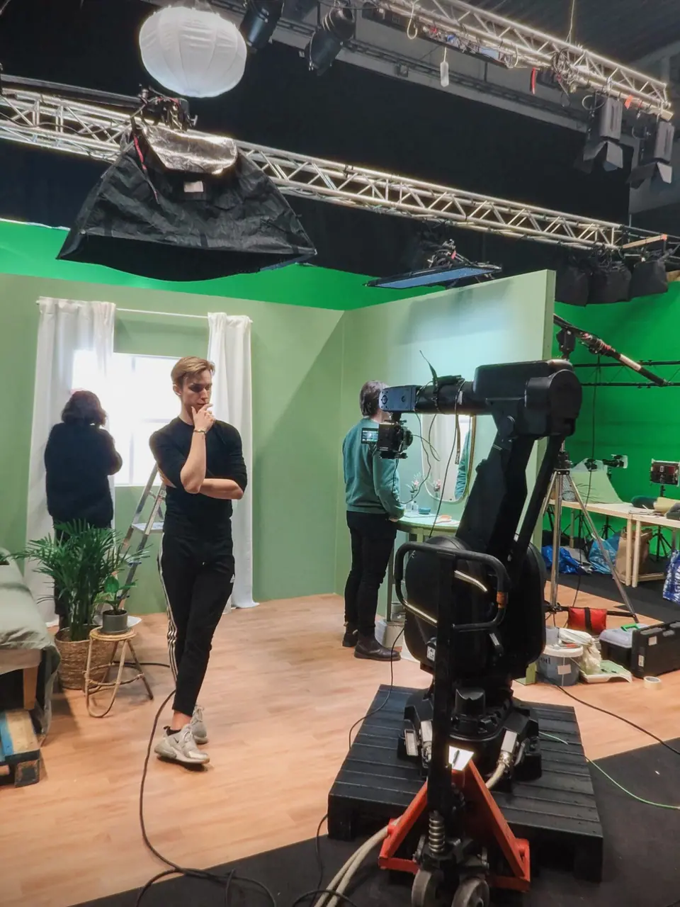 A camera on a robotic arm in front of a sage-green room set in a green-screen studio, a crew member looking on