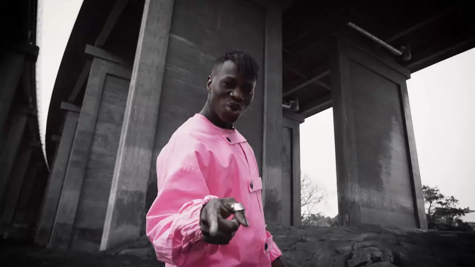 A man in a pink jacket pointing at the camera under a concrete bridge