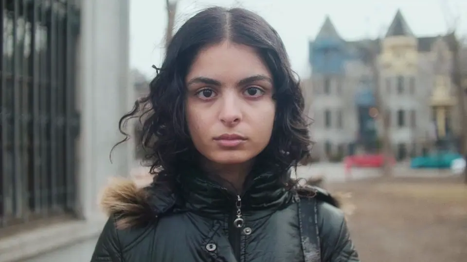 A young woman with long curly hair in a black puffer jacket, looking into the camera