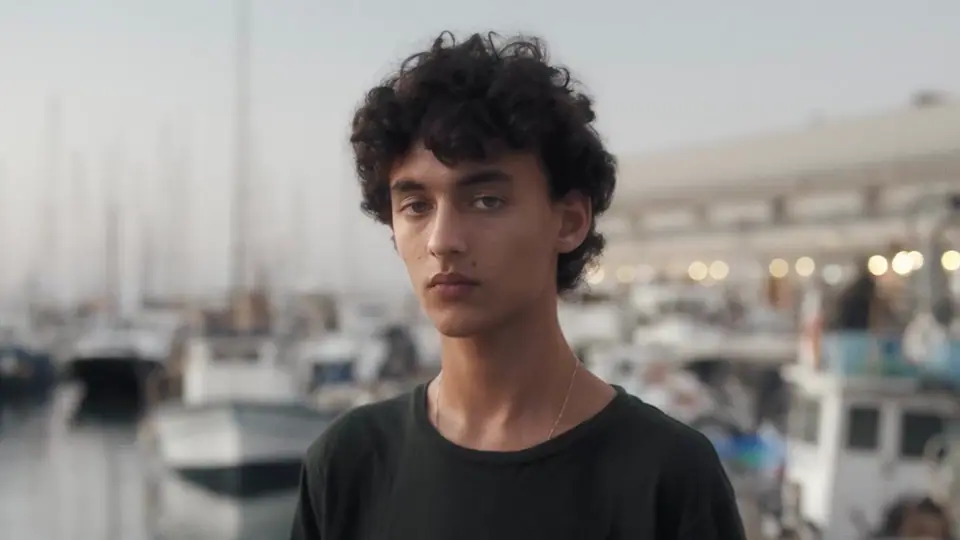 A young man with curly hair in a black T-shirt at a marina
