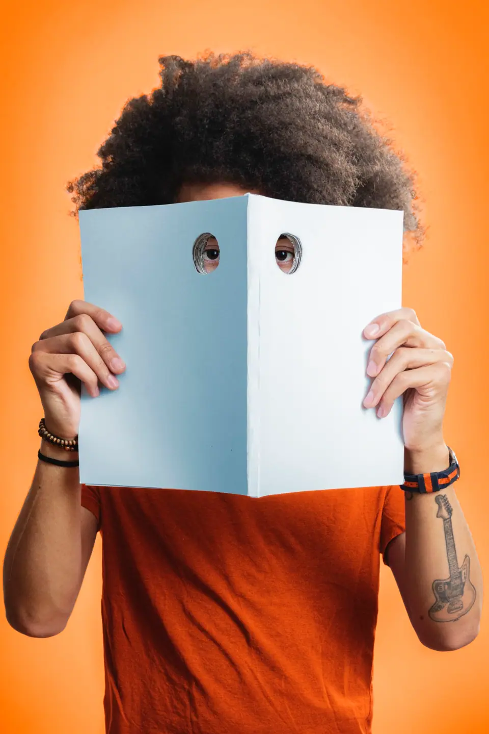 A man in an orange T-shirt peeking through two holes cut in an open book, against orange