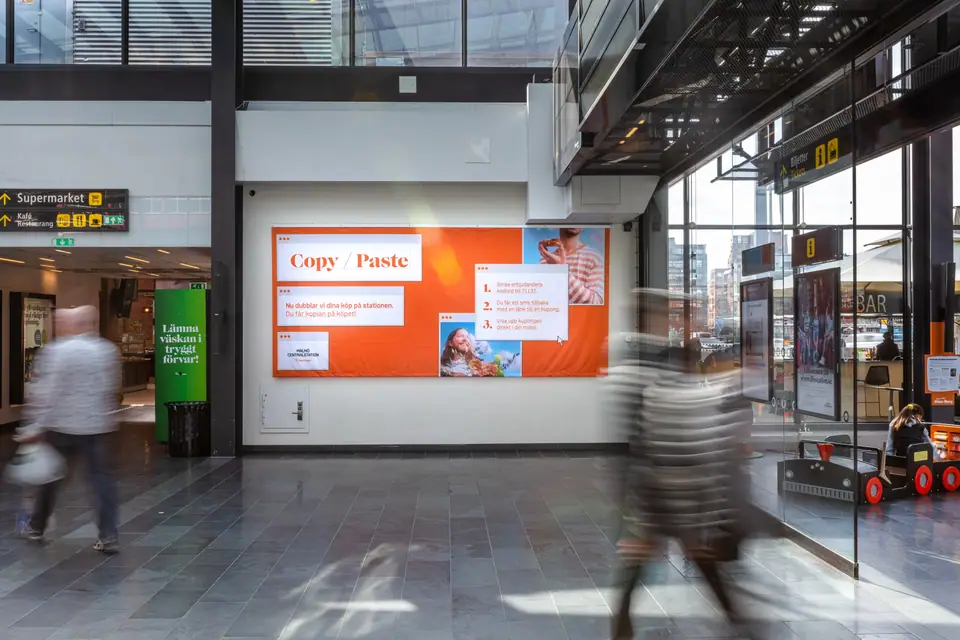 A large Copy/Paste campaign wall in a station hall, travellers blurred as they walk past