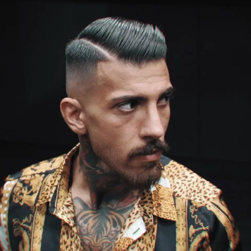 A tattooed man in a leopard-print shirt with a slicked side part, in profile against a dark background