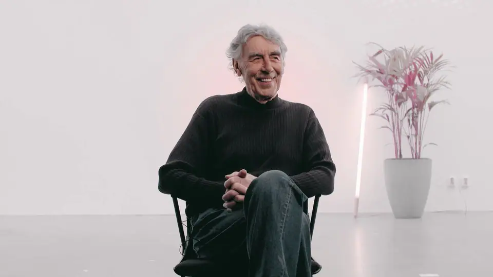 An older man in a black sweater smiling on a chair in a white studio, a pink plant beside him