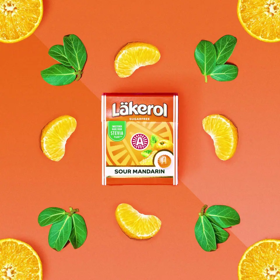 A pack of Läkerol Sour Mandarin surrounded by mandarin slices and leaves on orange