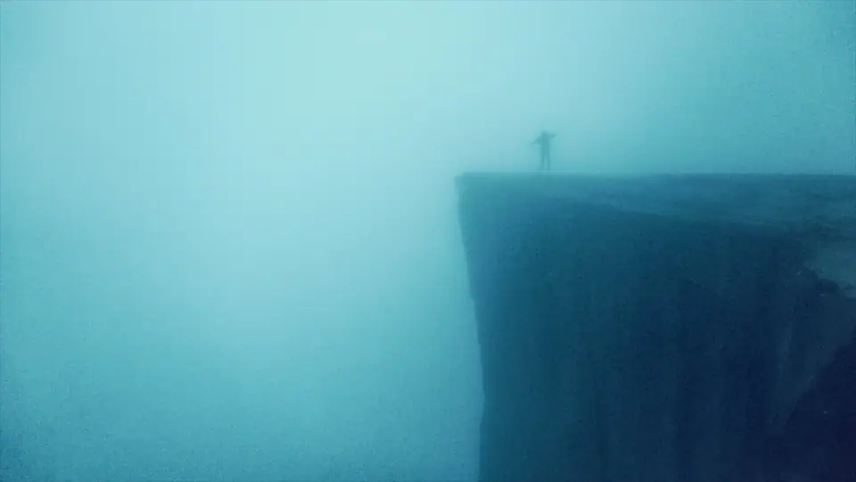 A lone figure standing at the edge of a cliff in thick blue fog