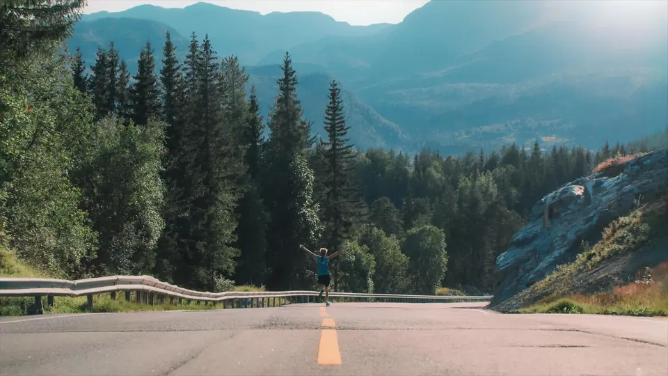 A man with his arms raised on an empty mountain road lined with pine trees