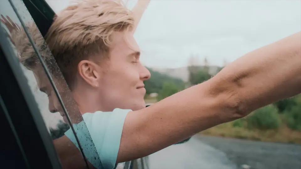A young man leaning out of a car window with his arm stretched out on a misty road