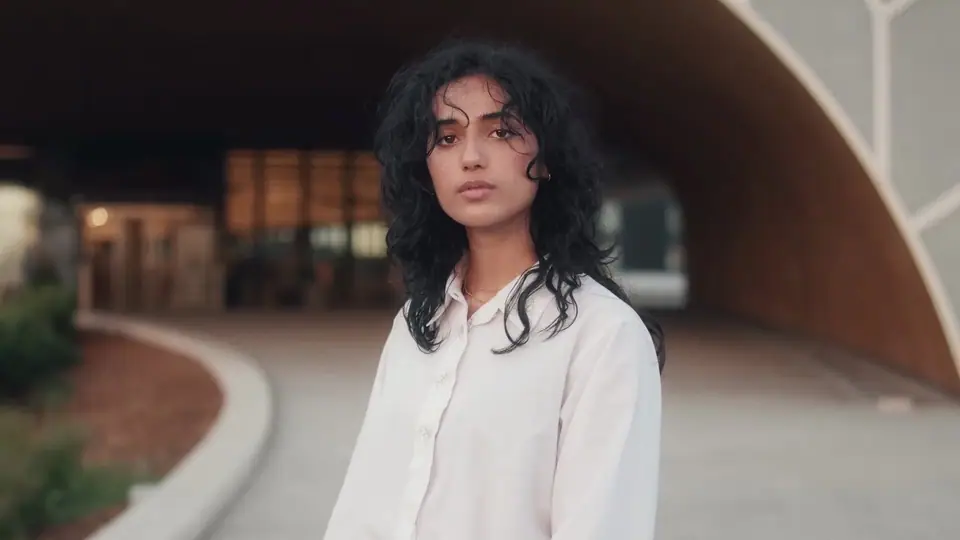 A young woman with long curly hair in a white shirt under a curved concrete bridge at dusk