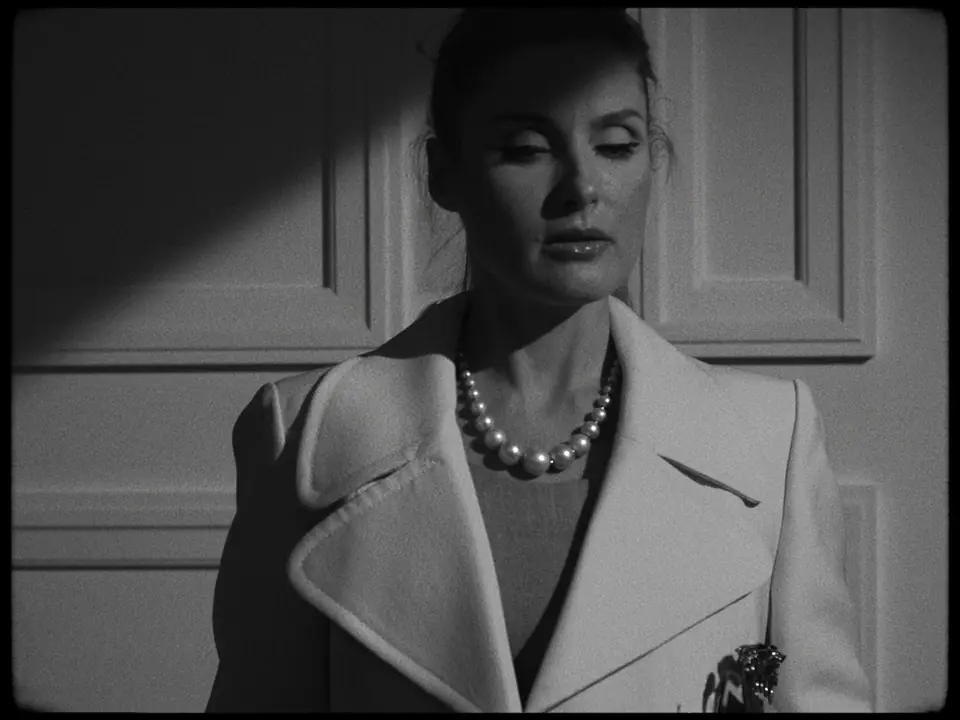 Black-and-white still of a woman in a white coat and pearls