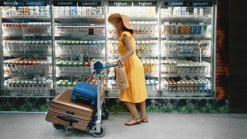 A shopper in a yellow dress and sun hat wheels a trolley with a suitcase past the dairy fridges